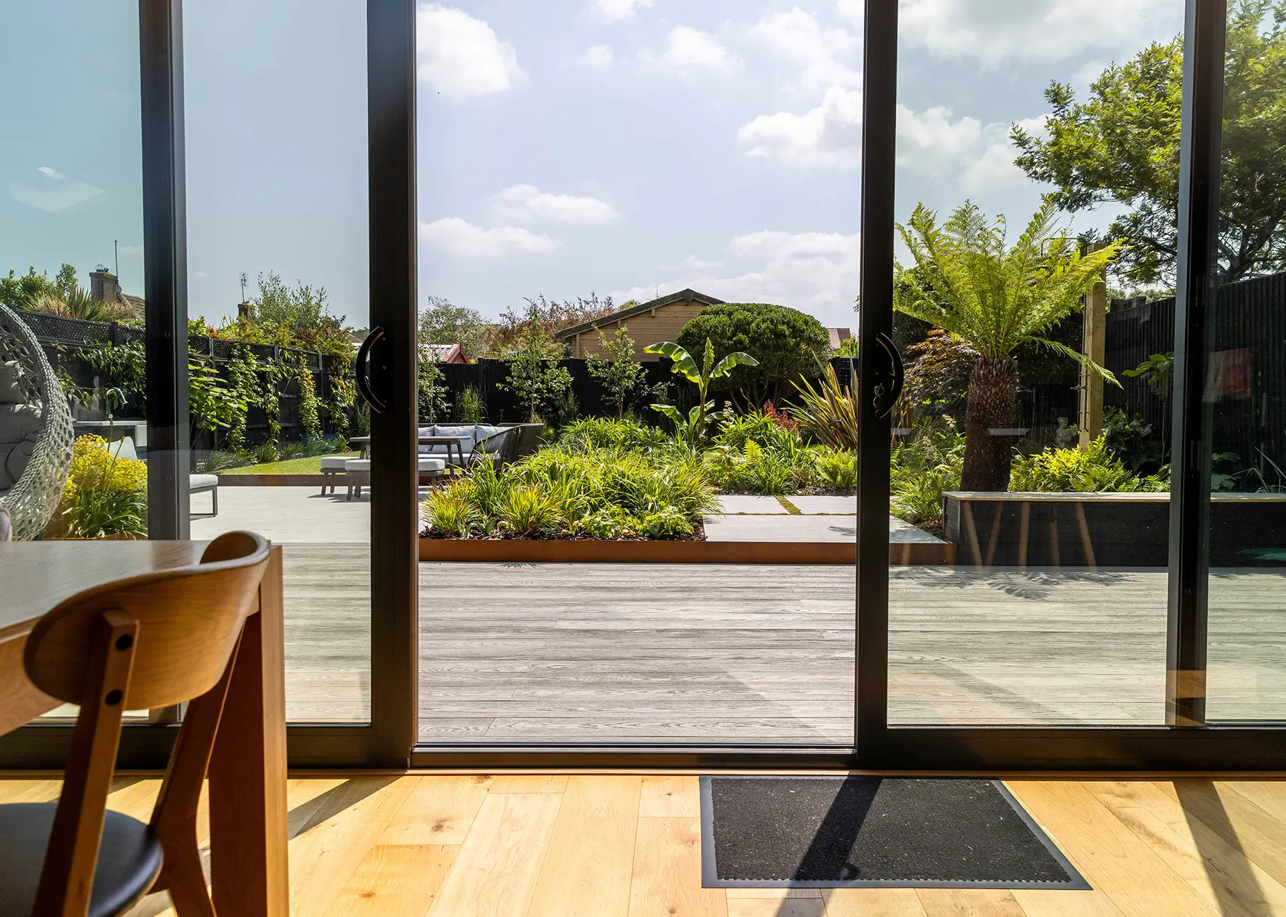 A view through sliding glass doors showcasing a green garden and bright blue sky beyond. A wooden chair is visible.