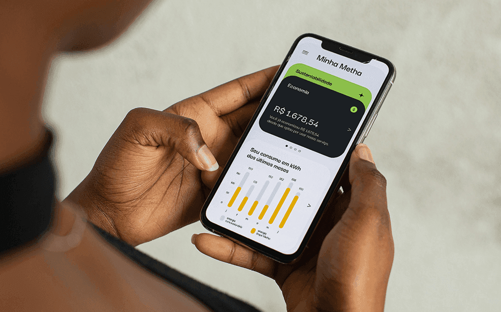 Mãos segurando um smartphone exibindo o aplicativo Minha Metha com gráfico de consumo de energia e valor economizado pelo cliente