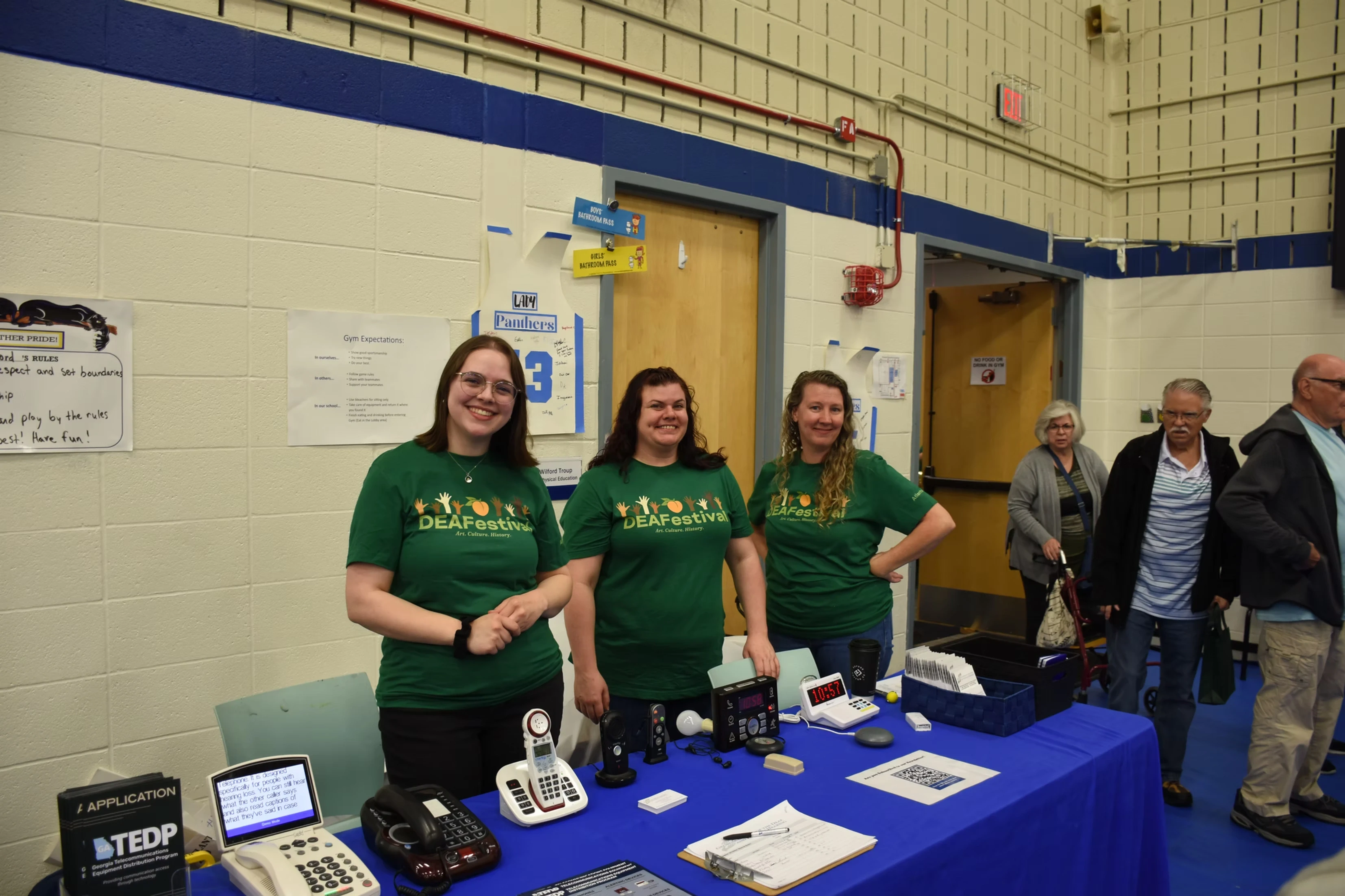 Three women smile at a table with assistive listening devices. They wear green DEAFestival shirts with a logo. People are in the background.