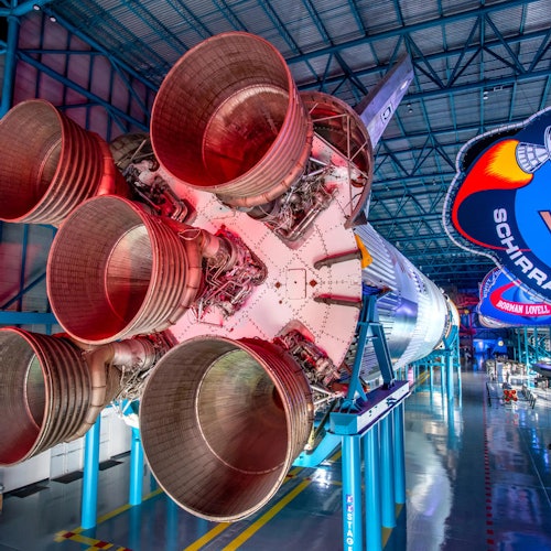 The image shows the large engines of a Saturn V rocket displayed indoors with signage and metallic structures surrounding it.