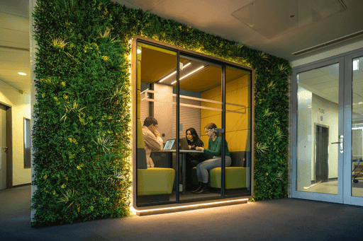 Drei Personen sitzen in einer modernen, von einer grünen Pflanzenwand umrahmten Besprechungsecke mit Glaswänden und hellgrünen Polstern