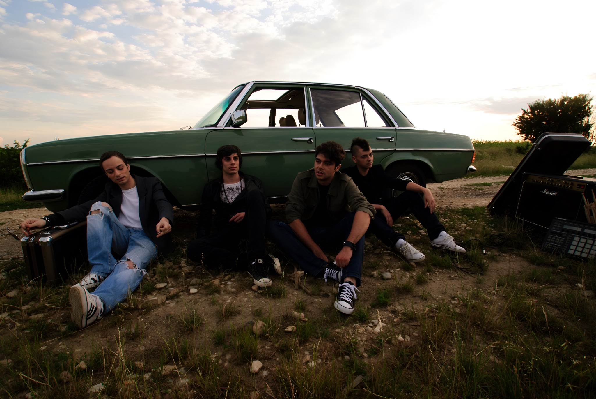 Plastik Charm band sitting on the ground near a vintage green car at sunset, with music gear scattered nearby.