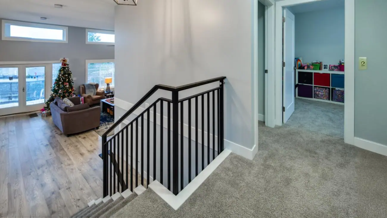 Upper floor landing in multi-level custom Burnaby home with elegant black metal staircase railing, plush carpeted hallway, dramatic view overlooking main living space, and access to children's play area