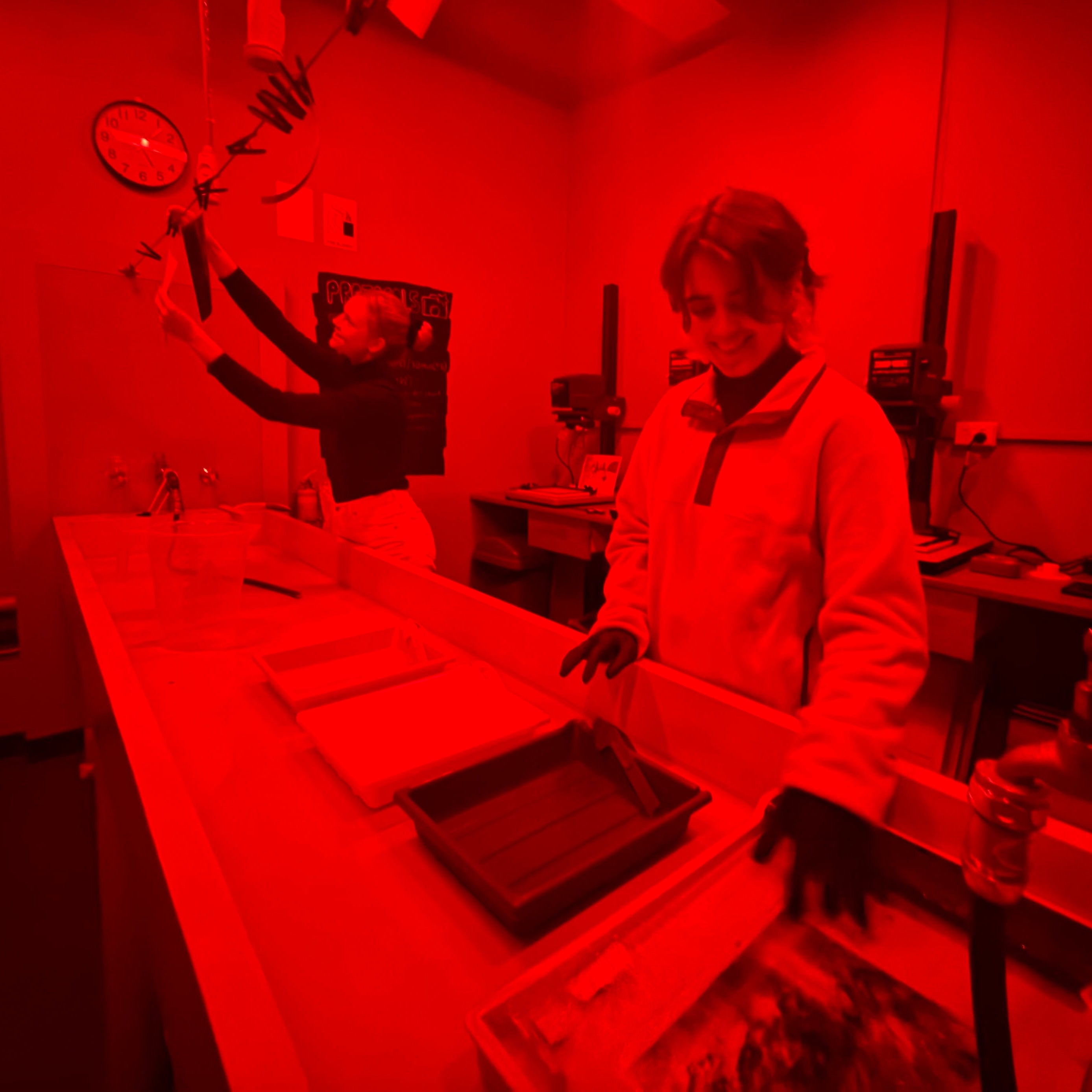 Person in a darkroom with red lighting, holding a camera and looking at prints on a light table.