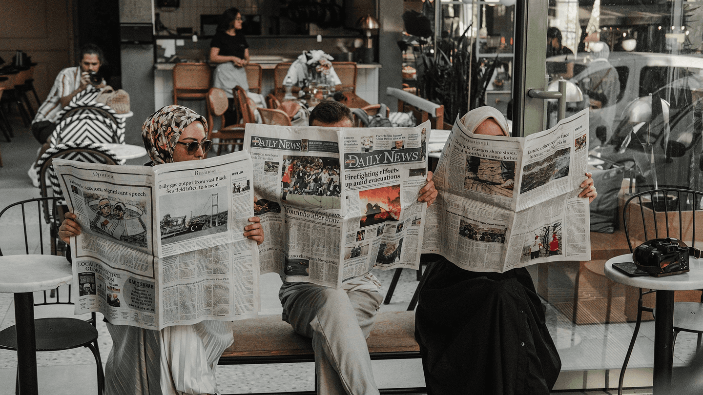 Several people sitting at a café table reading newspapers, with the papers covering most of their faces.