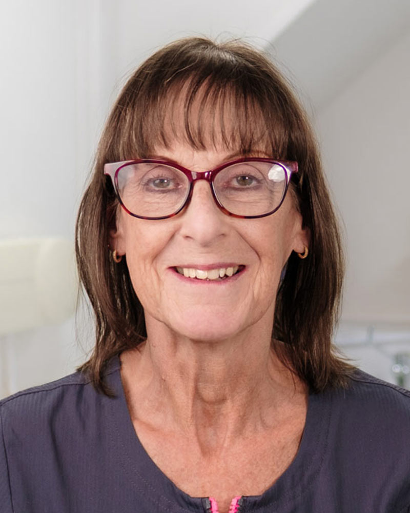 A portrait of Karen, a Dental Nurse at Cricklade Dental Practice, smiling and wearing a dark grey scrub top with a pink zipper pull and yellow embroidered text that reads "KAREN" on the left and "Cricklade Dental Practice" on the right. She is standing in a dental surgery with a dental light visible in the background.