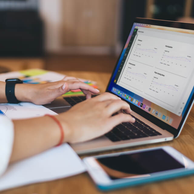 Person typing on a laptop displaying data analytics graphs, with a smartphone and notebook nearby on a wooden desk.