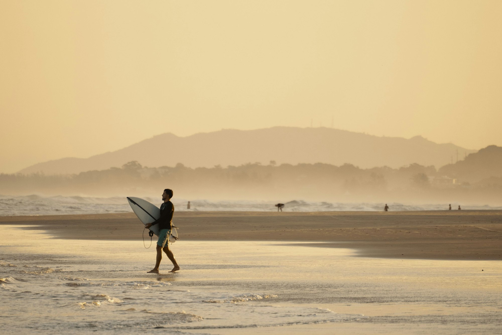 Surfer walks on beach at sunset with mountains