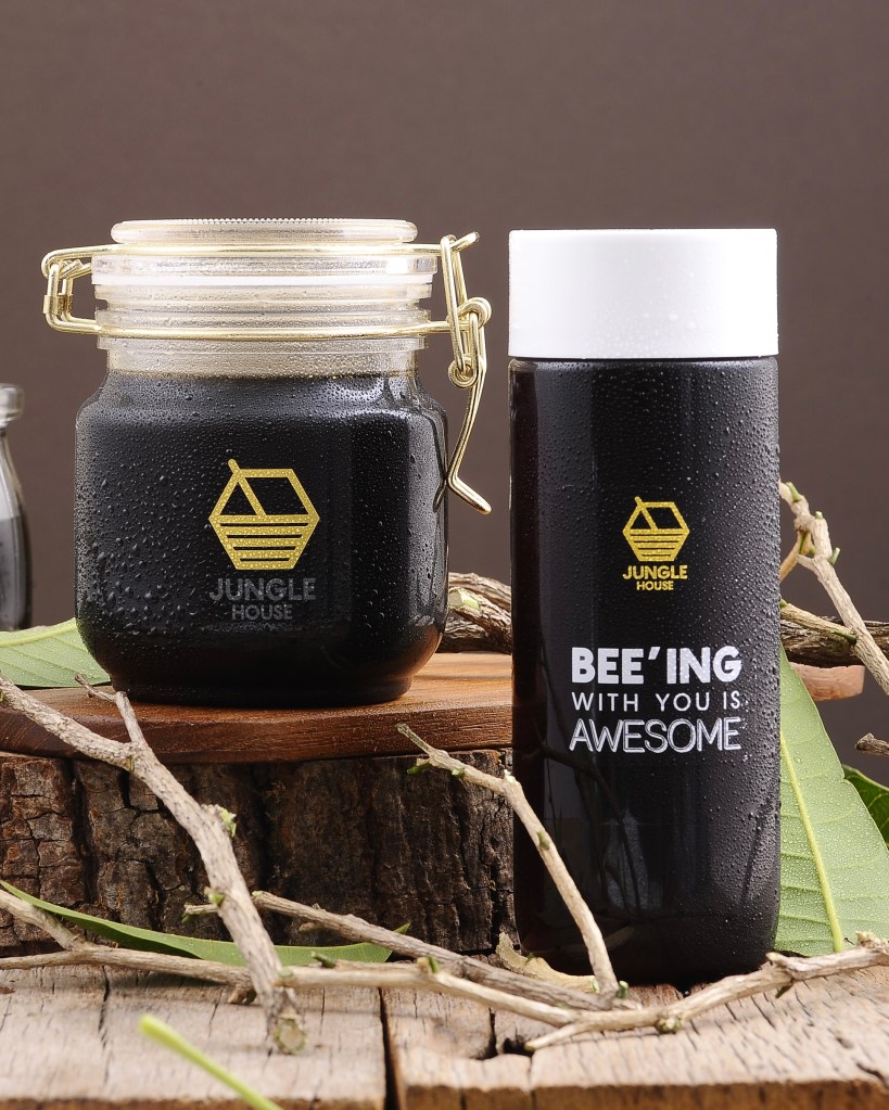 A glass jar and a plastic bottle of dark syrup, both featuring the 'Jungle House' logo. The jar has a gold-colored lid and is placed on a wooden surface with natural elements like branches and leaves.