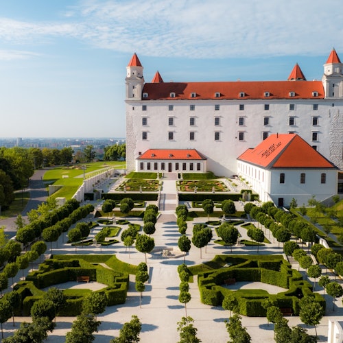 Un castillo con techo rojo y varias torres domina un jardín cuidadosamente arreglado con setos geométricos y caminos bordeados de árboles.