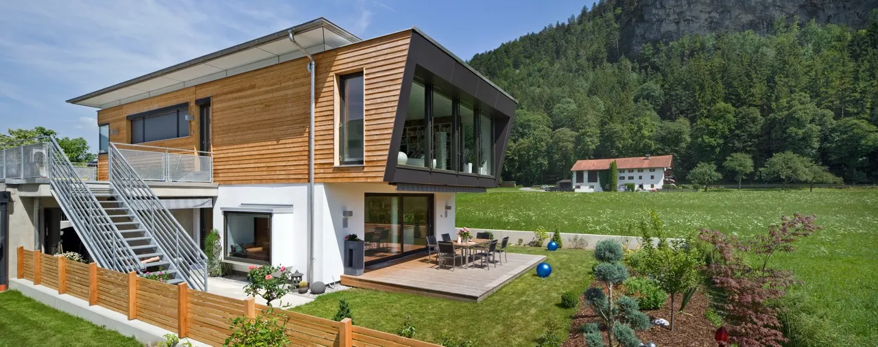 Modernes Holzfassadenhaus mit Terrasse und Garten, im Hintergrund bewaldete Berge und ein Bauernhaus.