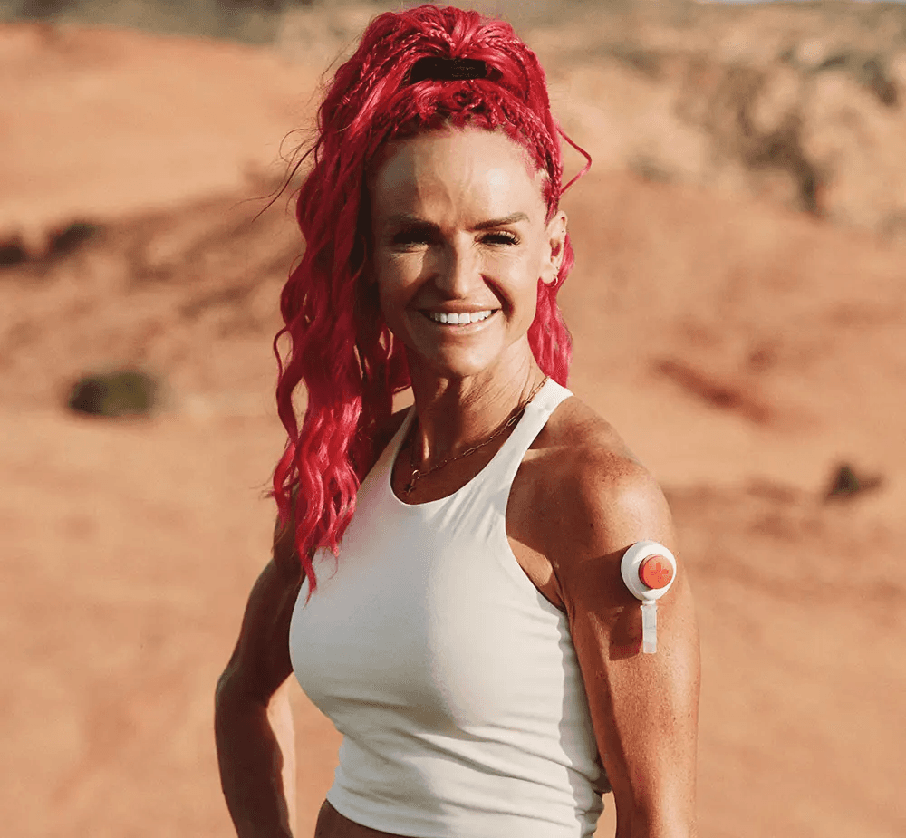 Fit woman with bright pink hair tied back, wearing a white athletic top, smiling outdoors in a desert landscape with a Rythm Health wearable sensor attached to her upper arm.