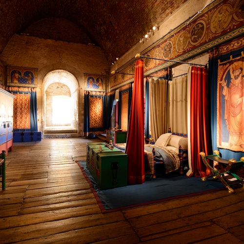 ห้องที่มีประวัติศาสตร์ พื้นไม้ประดับด้วยผ้าม่านสีสันสดใส ภาพปักผ้า เตียงคลุมด้วยหลังคา และเครื่องเรือนสไตล์ยุคกลาง