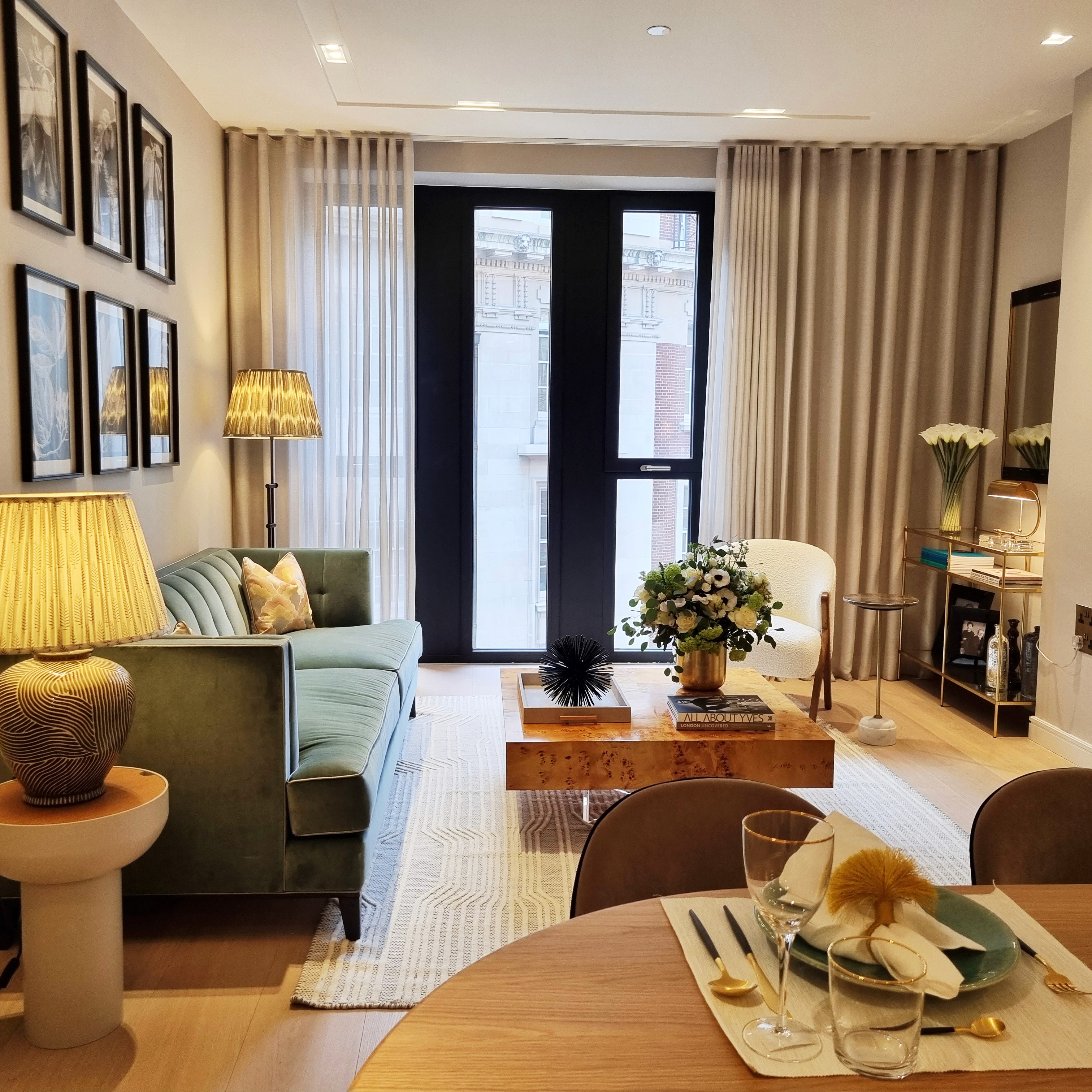 A modern, elegantly decorated living room features a plush green velvet sofa, a wooden coffee table adorned with a floral centerpiece, a stylish lamp, framed artwork on the walls, sheer curtains, and a round dining table with place settings, all set against a backdrop of large windows.