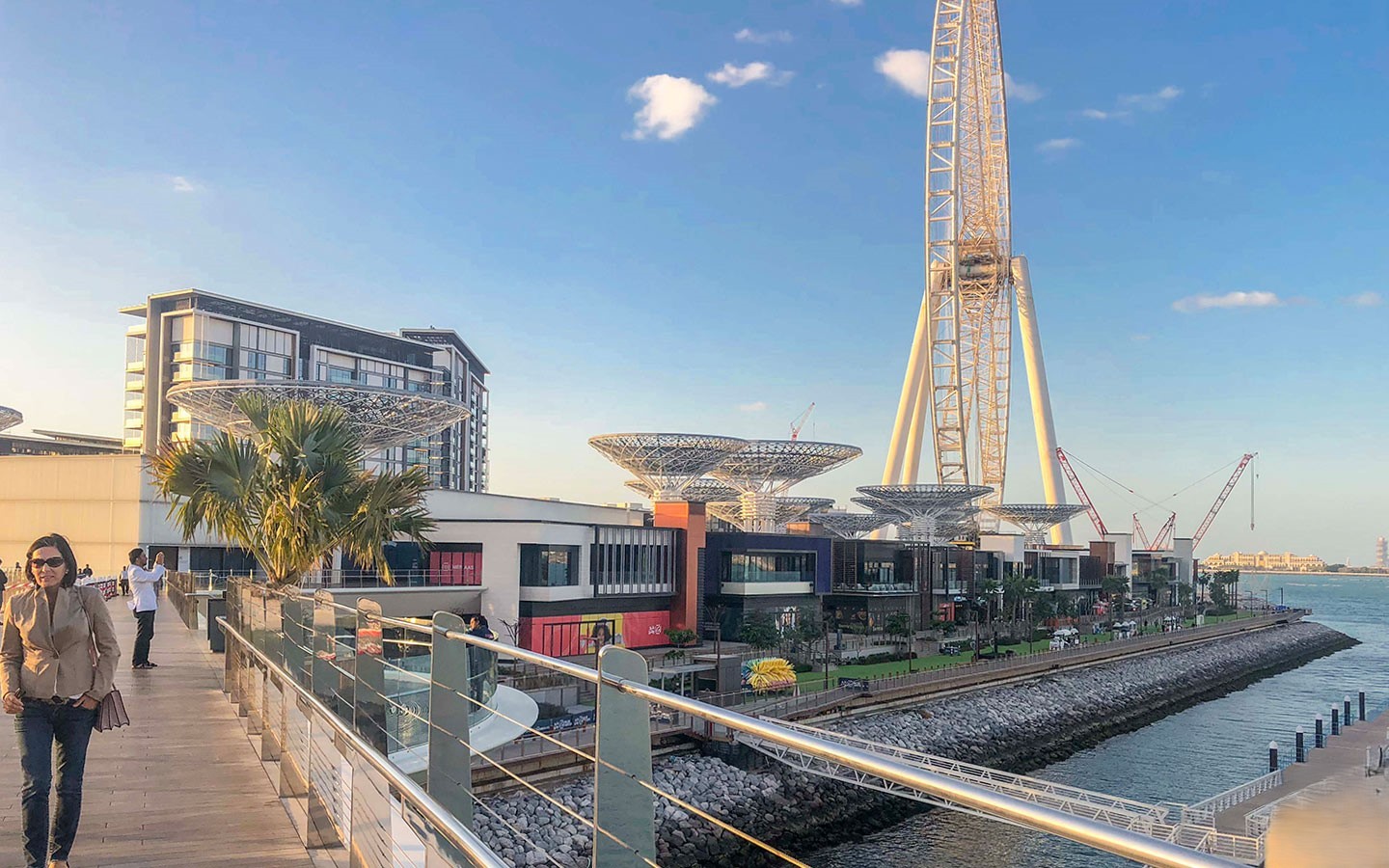 A pedestrian walkway along the water on Bluewaters Island, with low-rise retail buildings, a hotel, and the Ain Dubai Ferris wheel structure in the background.