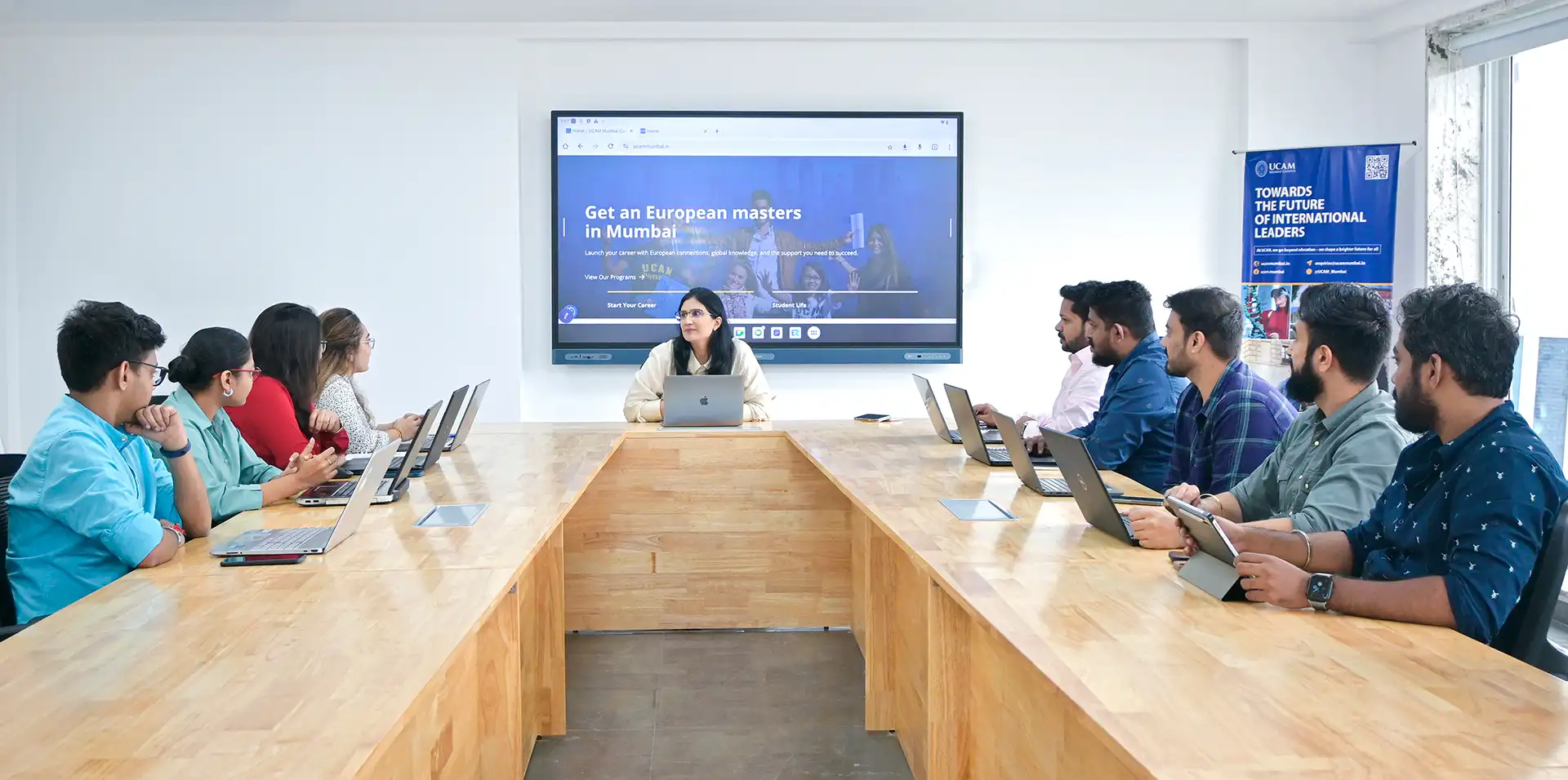A group of people sitting around a large wooden conference table, engaged in a meeting with laptops open, while a presentation on "European Masters in Mumbai" is displayed on a screen in a bright, modern room at European University UCAM.