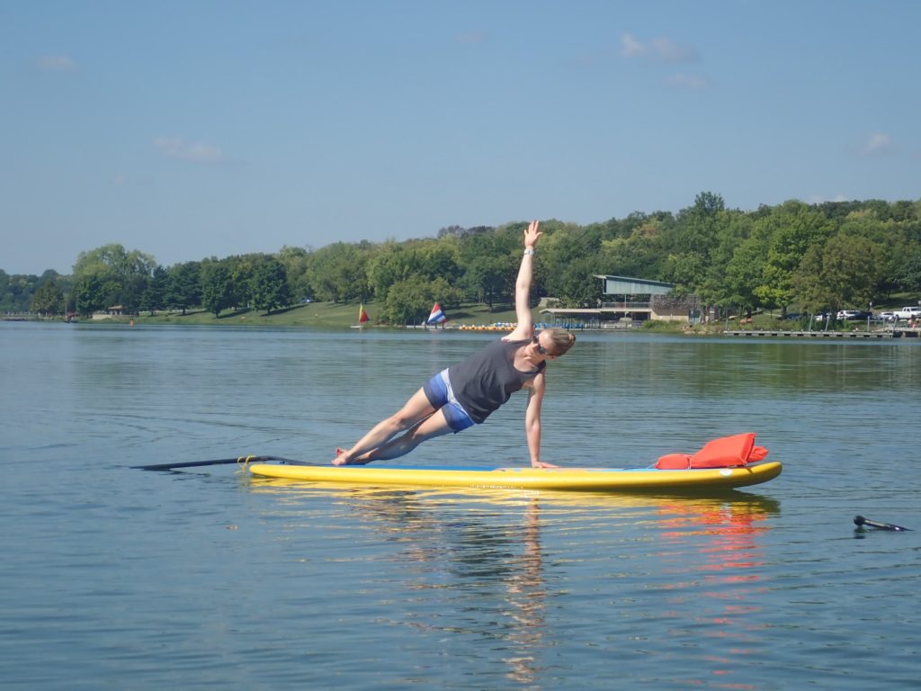 Heather Hausman performs a side plank yoga pose on a yellow paddleboard, set against a calm lake with a tree-lined shore under a clear blue sky.