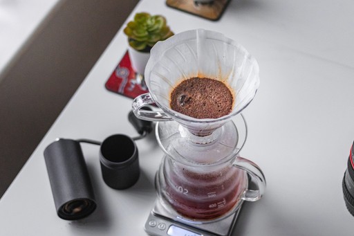 A top-down view of a Hario V60 filter with coffee being brewed into a Hario carafe. A succulent and a coffee grinder are also visible in the image.