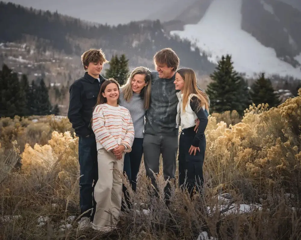 A happy family of five posing together in a brush-filled field during one of their Aspen Family Photo Shoots, with a snow-covered mountain slope in the background.