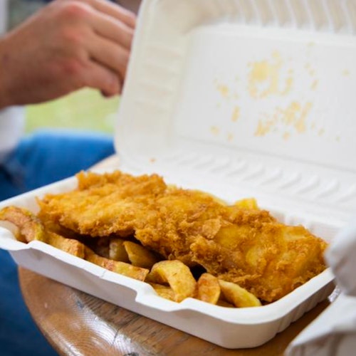 一个人在户外座位上拿着一个泡沫塑料盒，里面装着炸鱼和薯条。