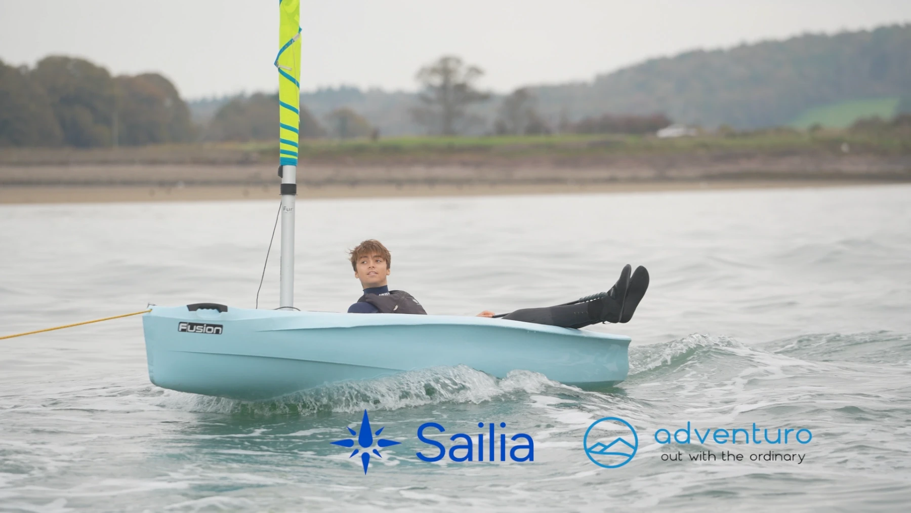 A young person sails a small, blue sailboat with a colorful mast on calm waters. The shore, with trees and hills, is in the background. Logos for "Sailia" and "adventuro" are in the corners, adding a sense of branding and adventure to the image.