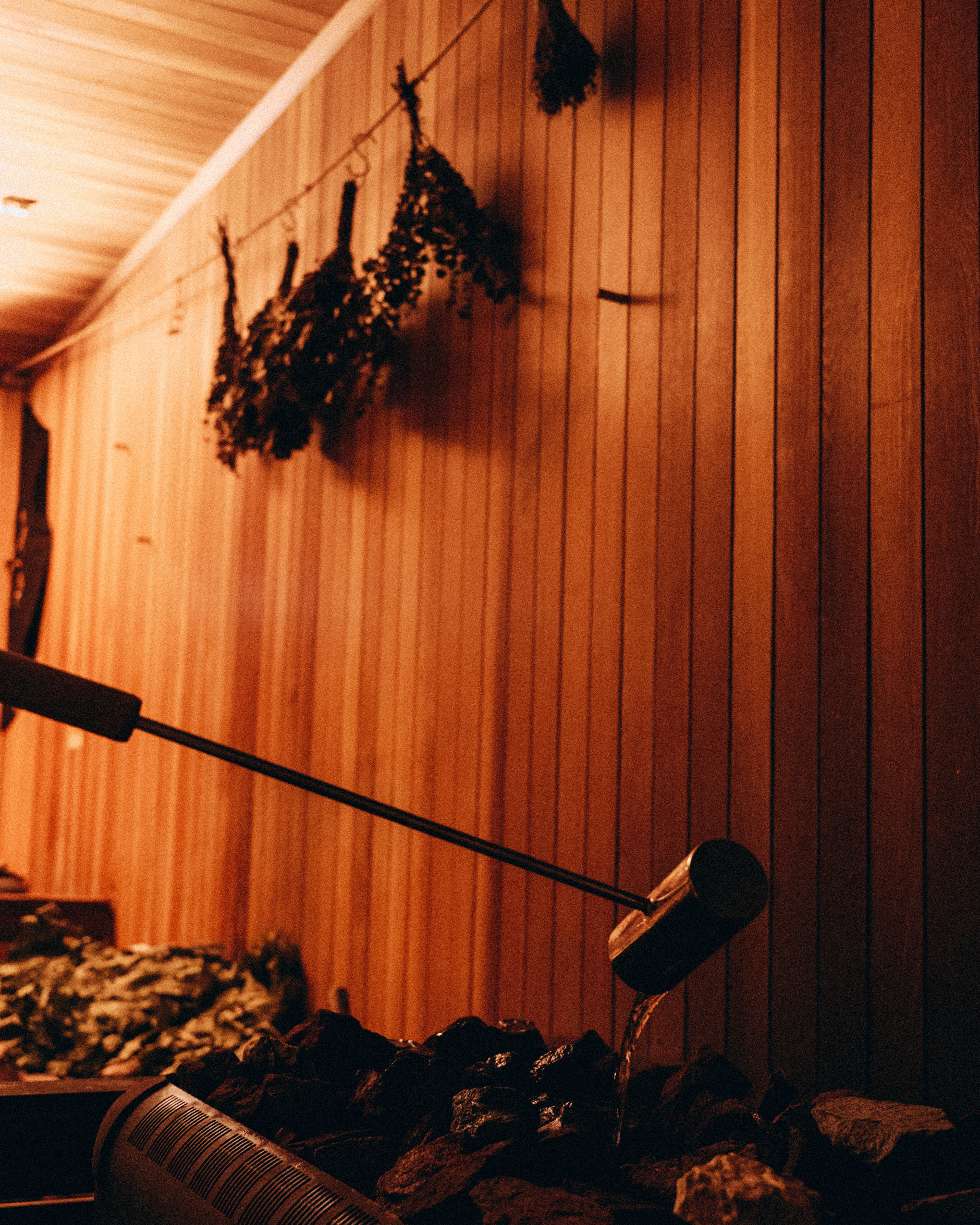 Wooden ladle pouring water onto hot sauna stones beneath bundles of birch leaves hanging on the wall
