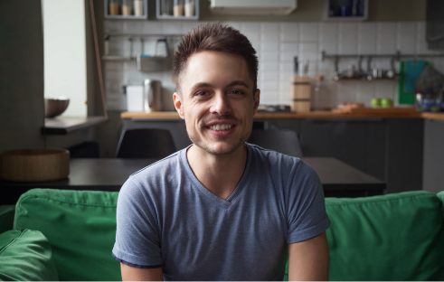 Smiling man on a video call, seated on a green sofa in a home kitchen, looking at the camera.
