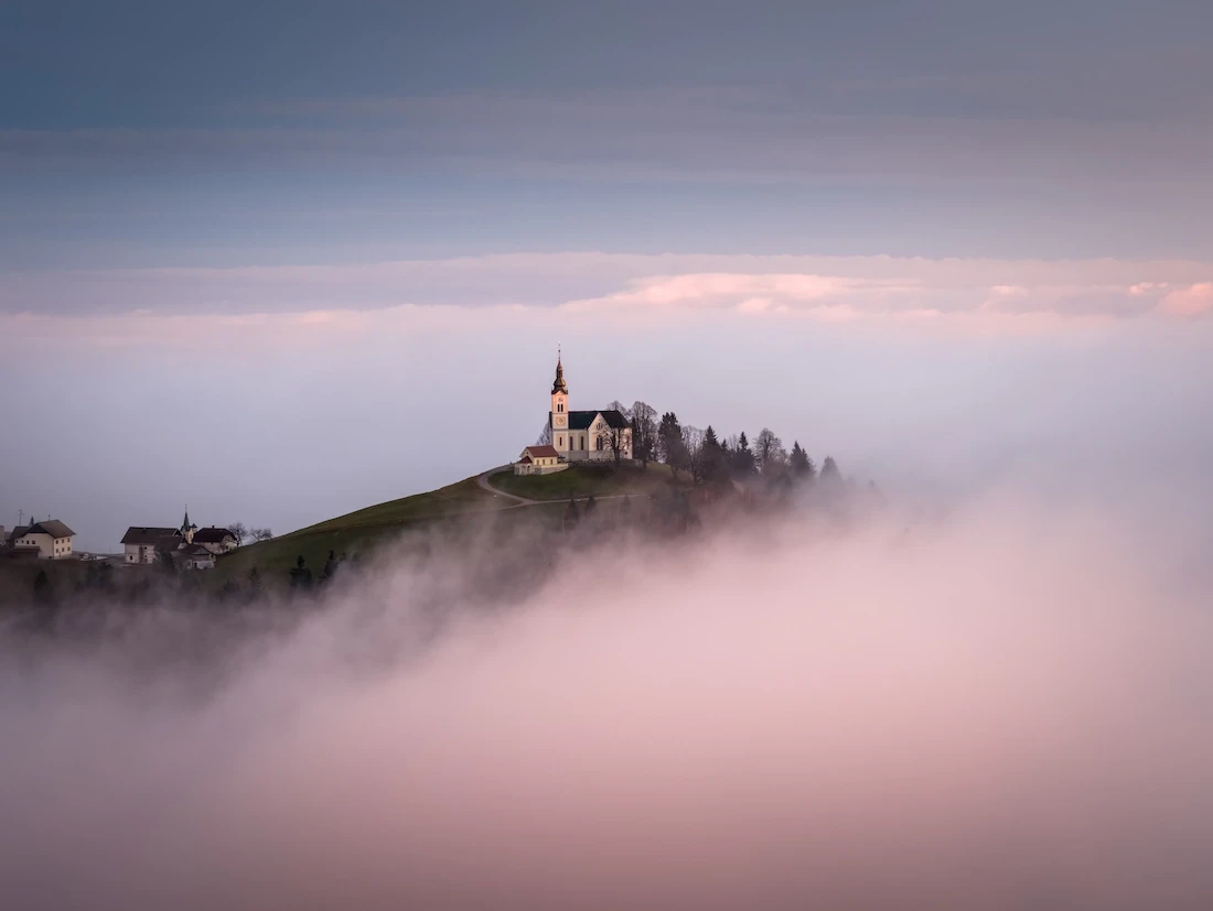 The iconic hilltop church of Sveto Lenart in Slovenia, appearing as an island above a dense layer of low-lying fog during a pastel sunrise.
