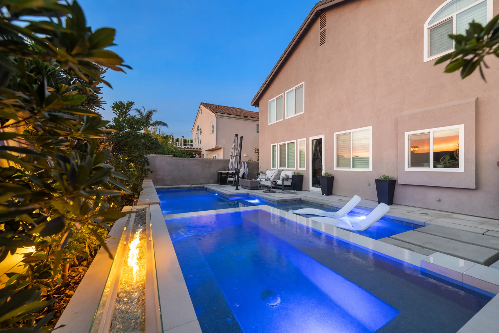 Illuminated pool with white loungers and a paved deck beside a home