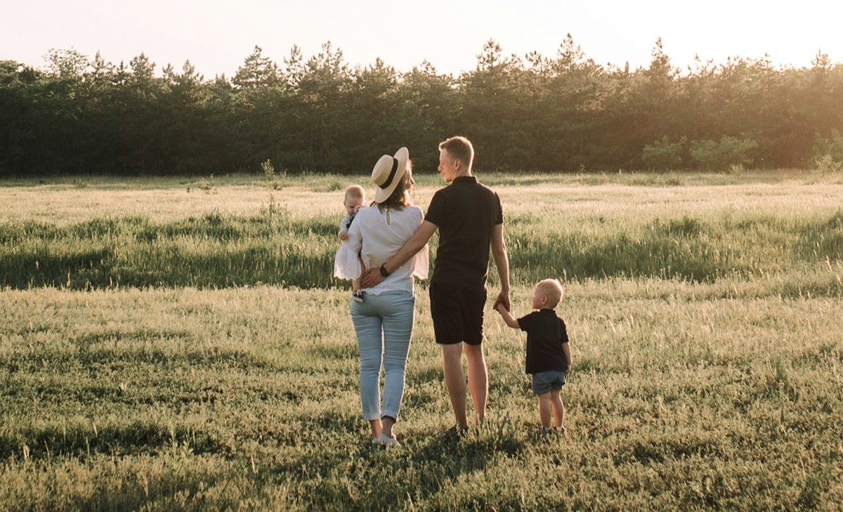 A family of four walks away from the camera through a field of tall grass toward a forest.