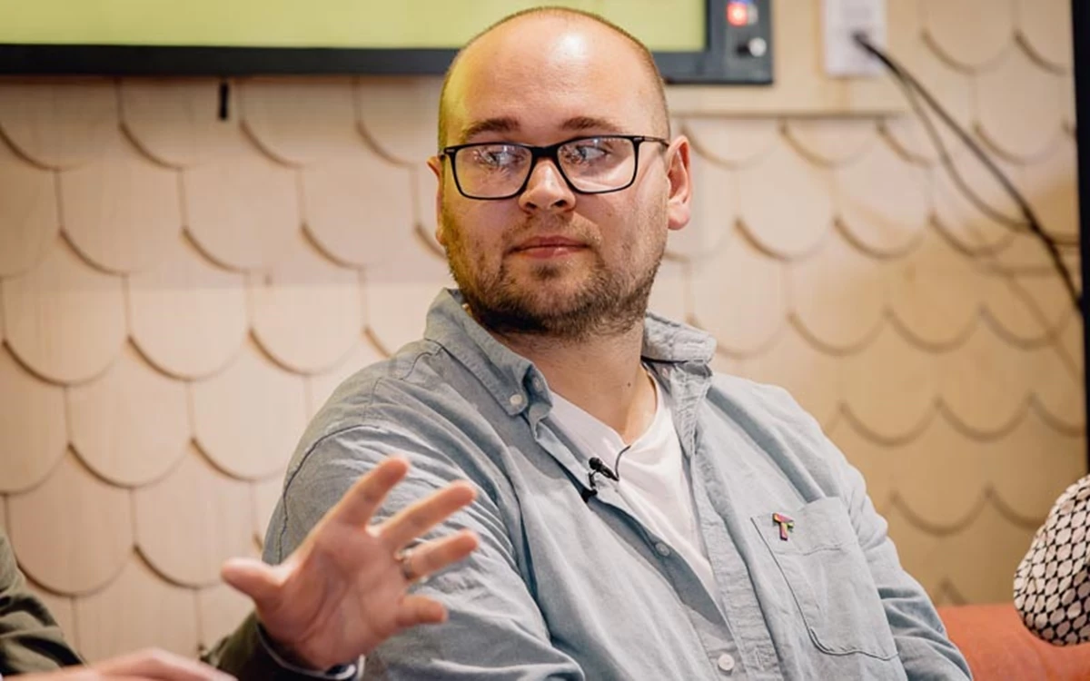 A panelist speaks while gesturing outward, seated in a relaxed stage setting. The image captures an active moment of explanation.