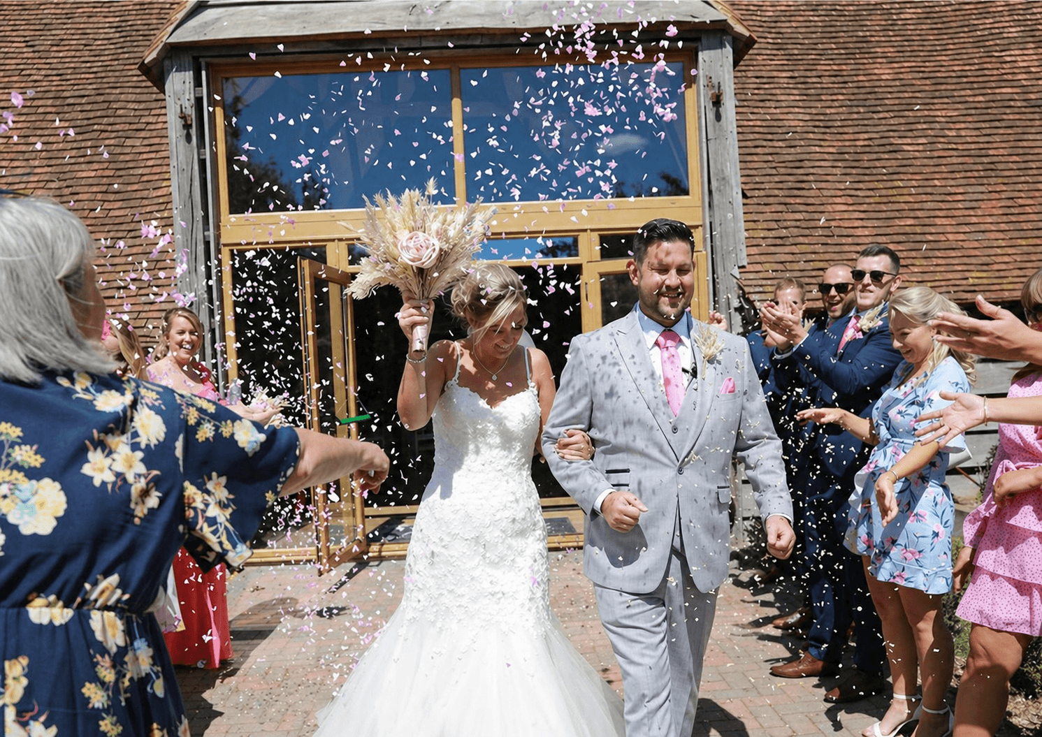 A smiling groom in a brown suit is embraced by his bride in a white,  lace wedding dress
