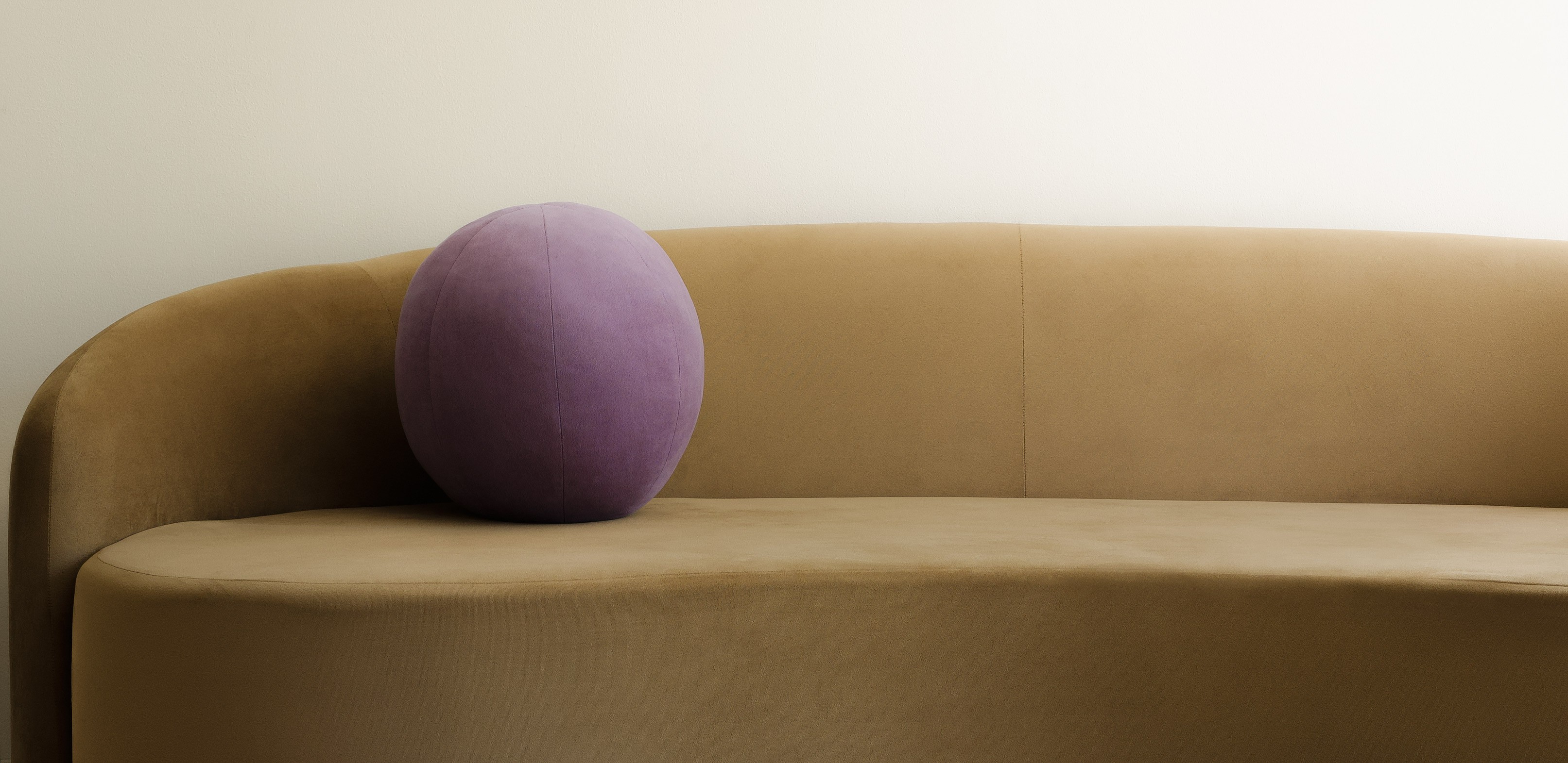 A minimalist scene with a beige curved sofa against a cream wall. A single purple sphere cushion adds a touch of color, creating a calm and modern atmosphere.