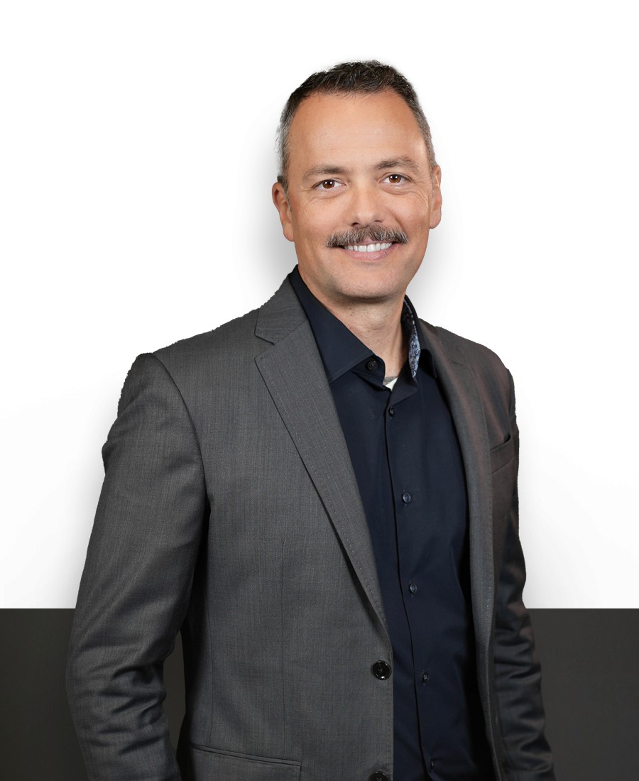 Professional headshot, man with light skin, dark hair and mustache, wearing a grey suit with navy collared shirt, relaxed and smiling.