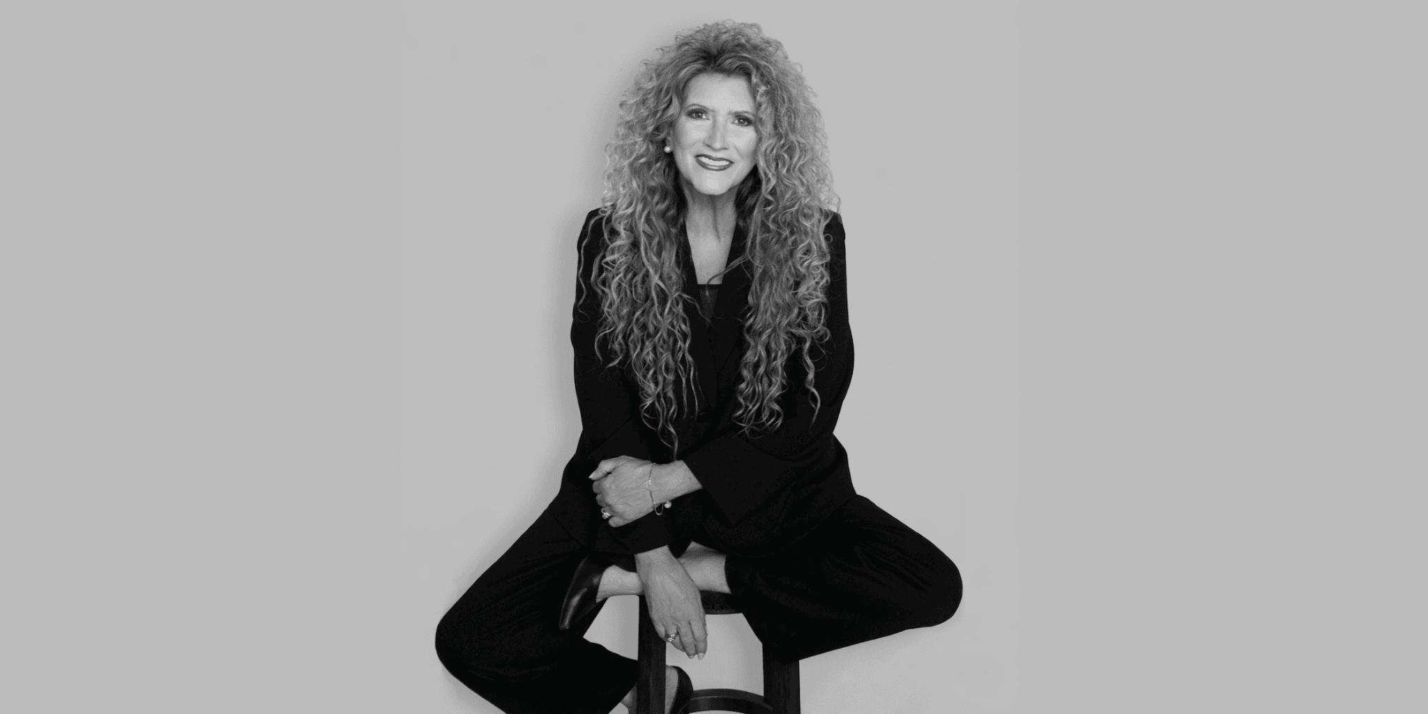 A woman with long, curly hair sits confidently on a stool in a black suit, smiling. The monochrome image exudes a professional and approachable aura.