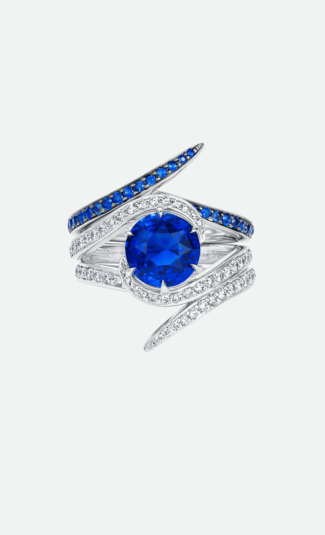 Close-up of an asymmetric multi-band ring with a round blue sapphire cradled by fluid, diamond-set white gold bands and a sweeping top crest of brilliant blue sapphires tapering to a sharp organic point.