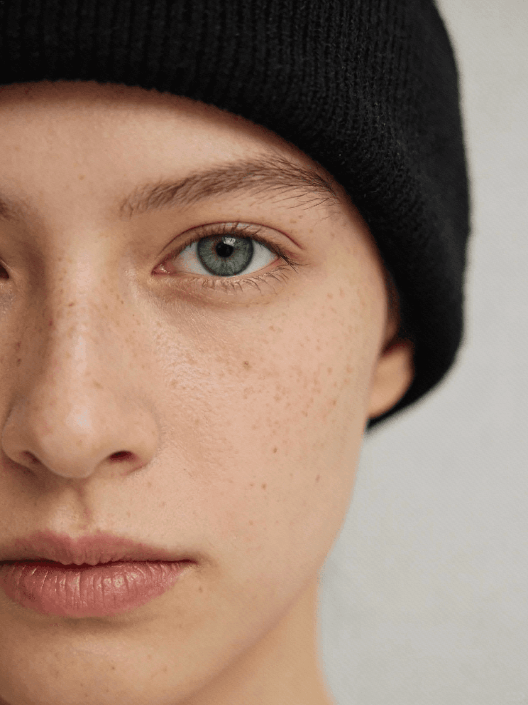 Close-up of a person's face with green eyes, freckles, and a black beanie, showcasing natural beauty and a neutral background.