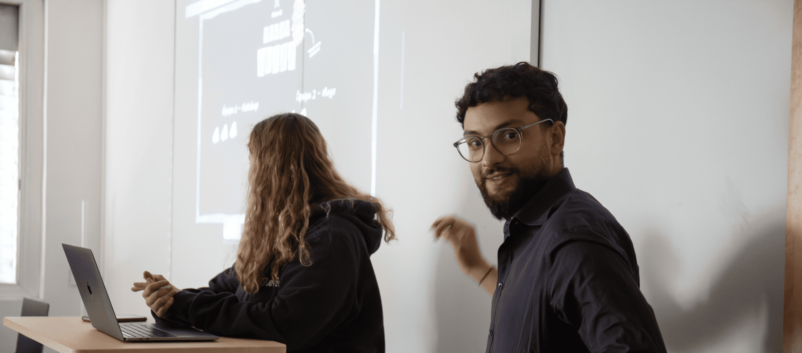 Homme en chemise blanche qui présente devant un tableau avec des post-it colorés face à une audience assise l’écoutant.