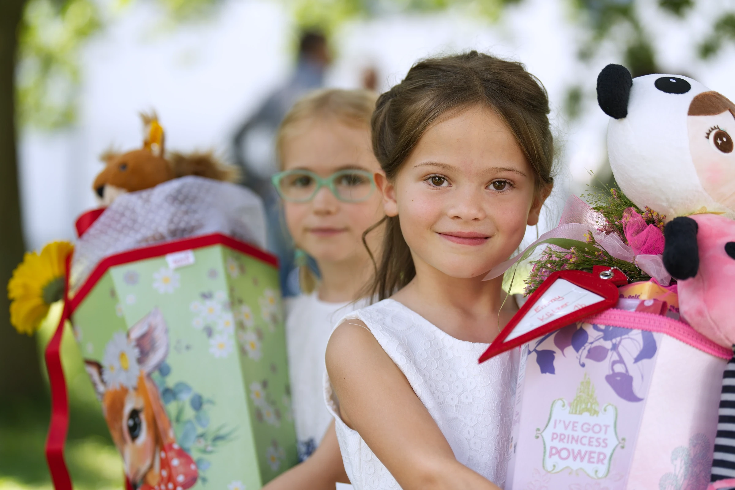 Emmy hält ihre Schultüte am Tag ihrer Einschulung und blickt lächelnd in die Kamera, während ein weiteres Mädchen im Hintergrund, mit zufriedenem Blick, ihre Schultüte hält und ebenfalls in die Kamera blickt.
