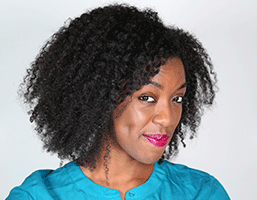 A close-up headshot of a woman with dark, curly hair. She wears a teal or turquoise crew-neck shirt and looks directly at the camera with a slight smile. Her lips are painted a dark pink or magenta color. The background is a plain, light gray or off-white.
