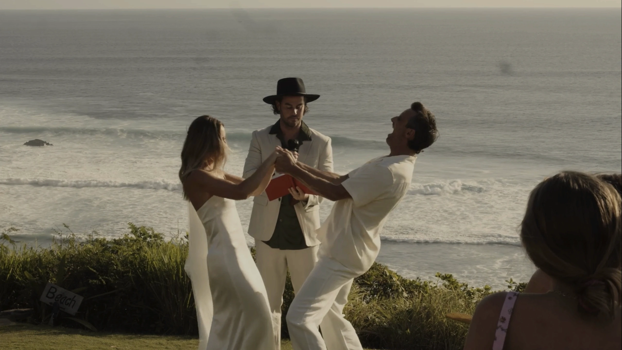 A couple in elegant white attire joyfully dances at an outdoor wedding ceremony on a grassy cliff overlooking the ocean, with a person in a light suit and black hat officiating under the clear sky.