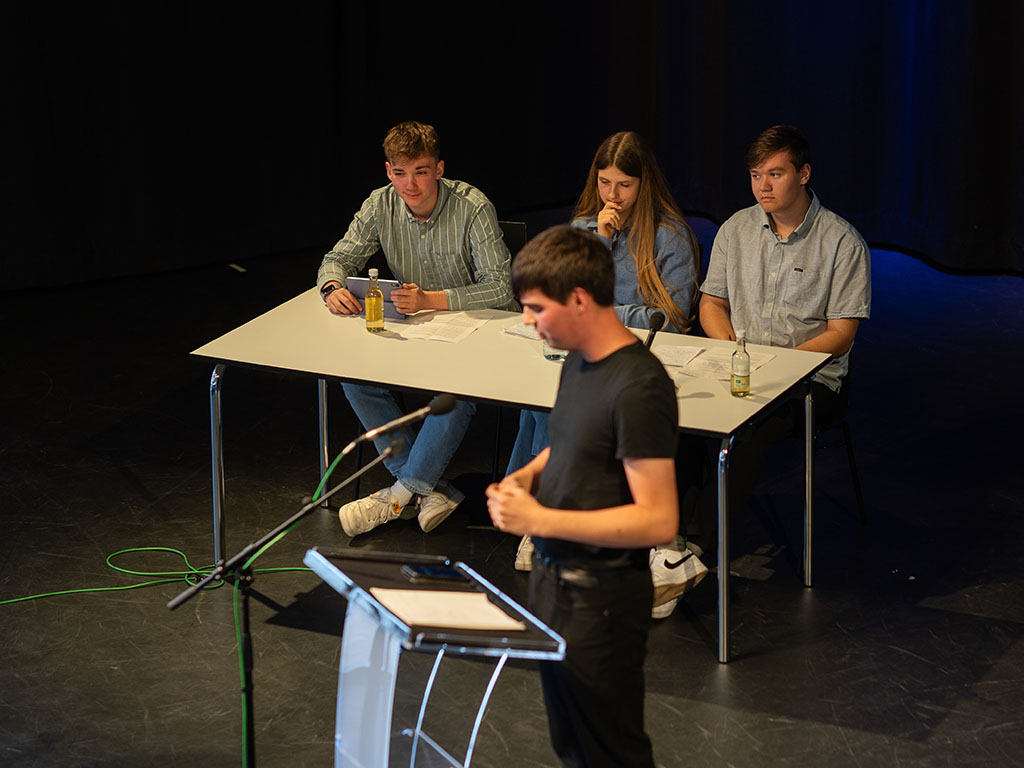 Jugendlicher Redner bei der Debattenschmiede diskutiert vor zwei Schülerteams auf der Bühne.