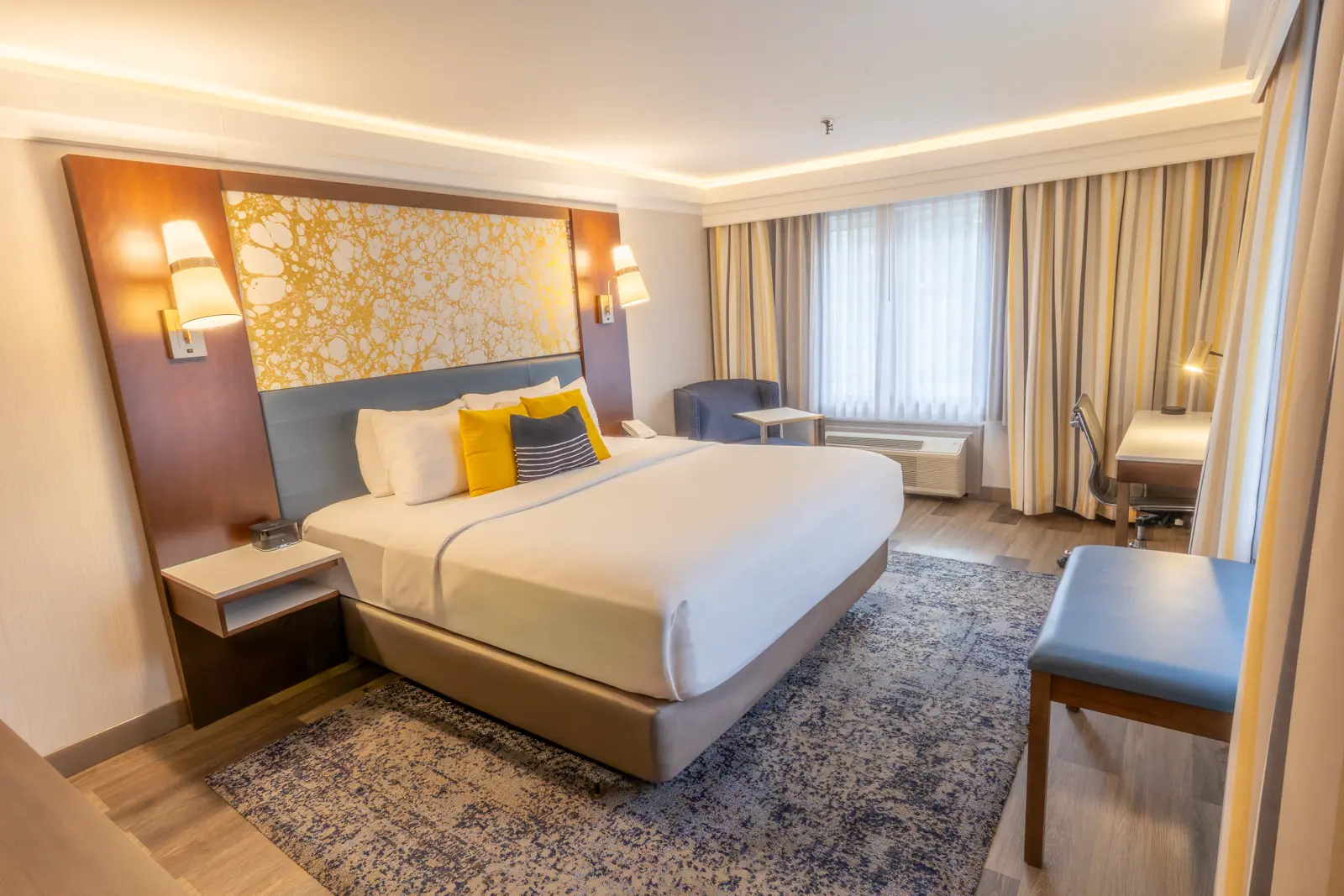 A modern hotel room with a king bed, gray sofa, and yellow chair.
