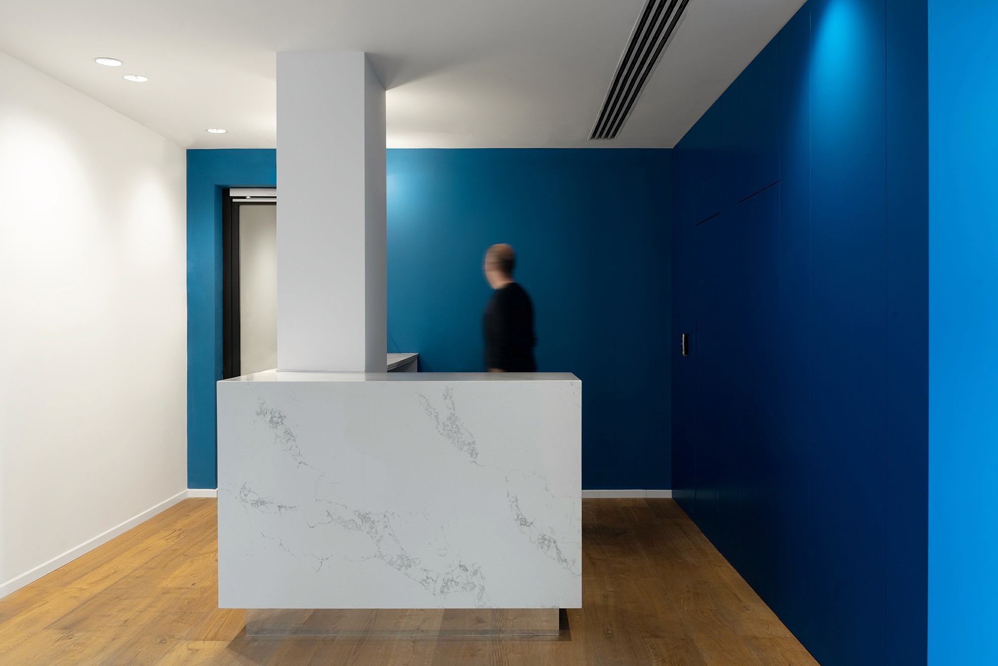 עיצוב כניסה: קוביית נגרות בגוון כחול עמוק המסתירה פונקציות שירות ומגדירה את אזור הקבלה, לצד חלונות רחבים לאור טבעי.