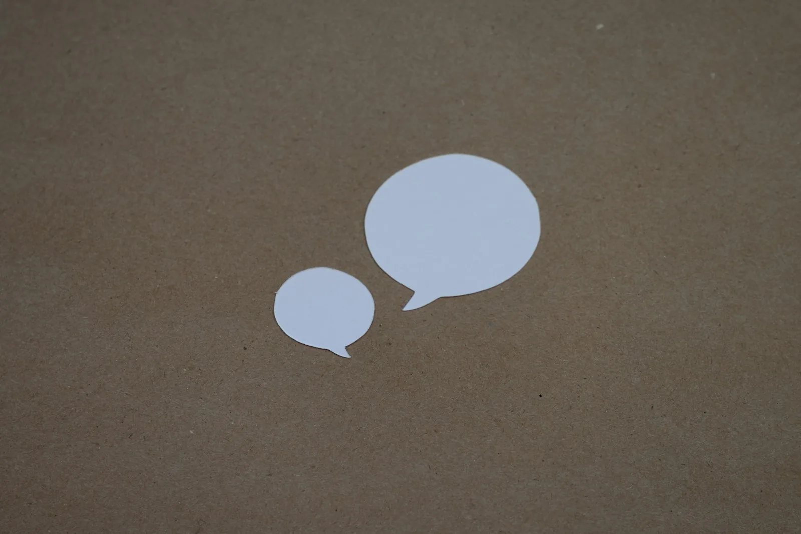 Two white paper speech bubble cutouts, one small and one large, lying on brown kraft paper
