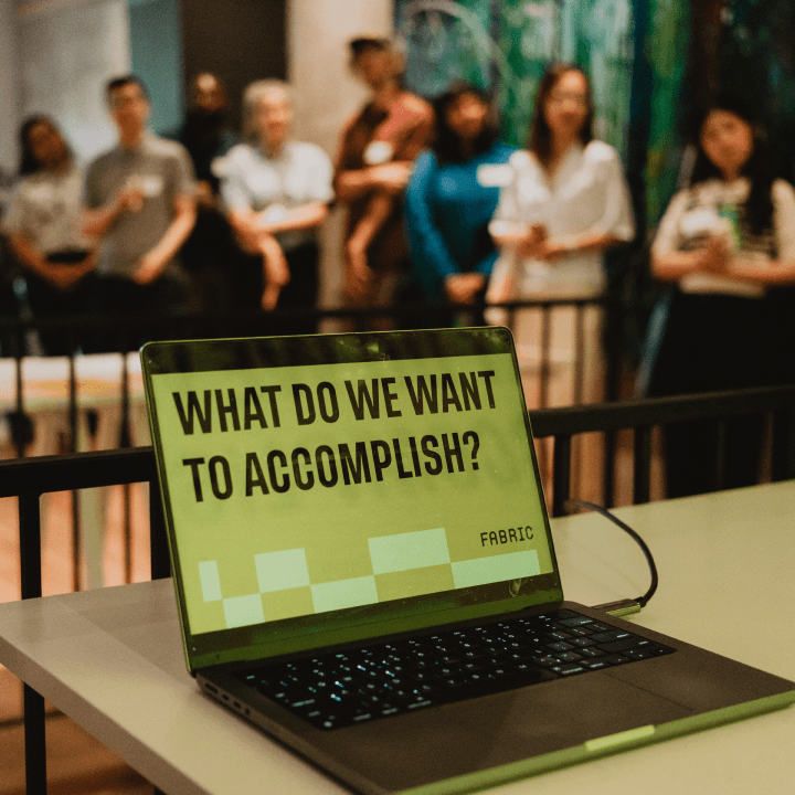 A laptop displays "What do we want to accomplish?" in bold text. A diverse group of people stands in the background, suggesting a meeting or workshop.