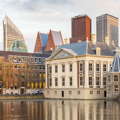 Un edificio histórico con un estanque en primer plano y modernos rascacielos al fondo durante el atardecer.