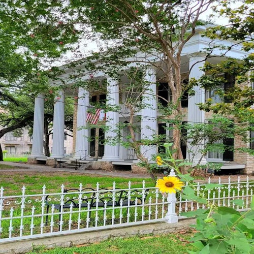 Edificio storico con alte colonne bianche e una bandiera americana, circondato da alberi e una recinzione in ferro battuto bianco con un girasole davanti.