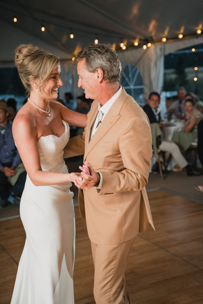 The bride laughing while dancing with a man (likely her father) in a tan suit under a wedding tent. Both are smiling warmly at each other during the reception.