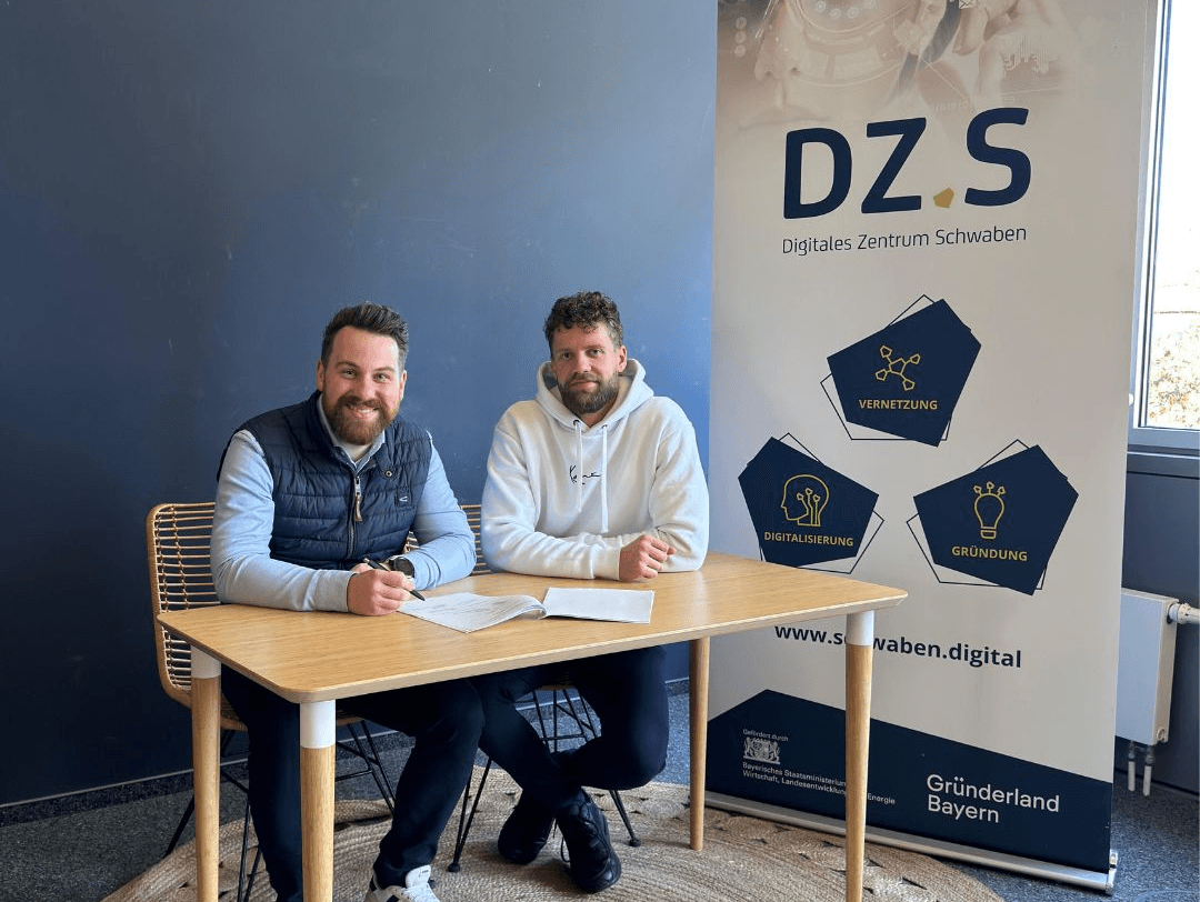 Two man sitting on a table, smiling to the camera and sign some documents. In the Background there is a rollup from DZ.S